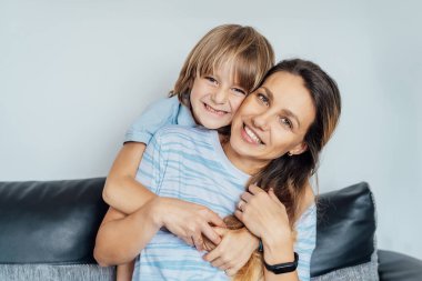 Evde gülümseyen mutlu bir ailenin portresi. Oğlan ve anne kanepede oturup kameraya bakıyorlar, aile hafta sonu boyunca birbirlerine sarılıyorlar. Seçici odak.