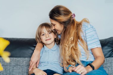 Evde gülümseyen mutlu bir ailenin portresi. Oğlu ve annesi kanepede oturuyor, birbirlerine sarılıyorlar, anne oğlunu öpüyor hafta sonu aile tatilinde. Seçici odak