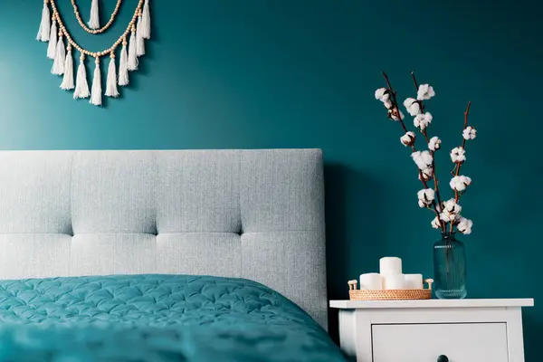 Stylish modern bedroom in dark colors. Cozy interior with blue turquoise walls, home decor. Bed with gray fabric headboard, blue blanket, bedside table, vase with natural cotton flowers, candles
