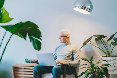 Orta yaşlı bir adam evde koltukta otururken dizüstü bilgisayar başında çalışırken yan bakıyor. Kendinden emin, tecrübeli, profesyonel bir erkek. Küçük işletme girişimcisi evde iş yönetir.