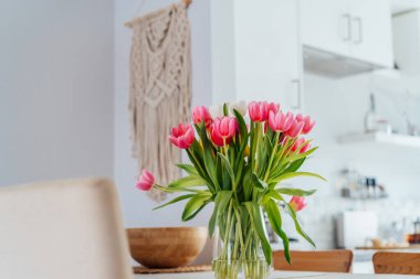 Şık ve modern boho, açık uzay beyaz mutfağının iç mekanı ahşap mutfak masasındaki vazoda pembe lale çiçekleri, duvarda makrome. Samimi Tasarım ev dekorasyonu. Seçici odak.