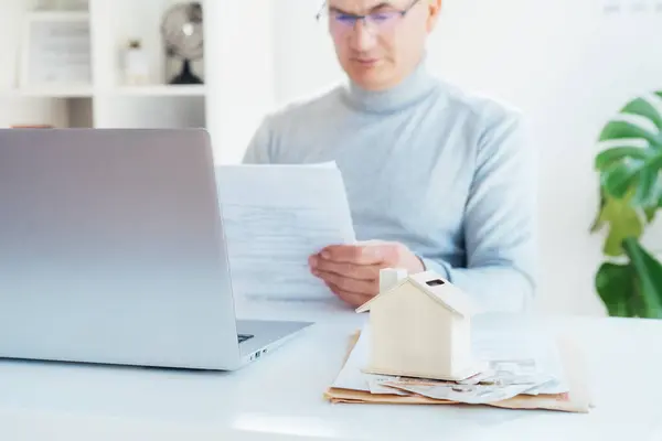 House model money box with cash on table and blurred man with docs works on laptop. Housing cost and payments. Real estate, property investment. Home mortgage loan rate. Saving money for retirement