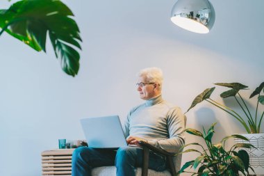 Orta yaşlı bir adam evde koltukta otururken dizüstü bilgisayar başında çalışırken yan bakıyor. Kendinden emin, tecrübeli, profesyonel bir erkek. Küçük işletme girişimcisi evde iş yönetir.