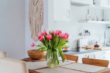 Şık ve modern boho, açık uzay beyaz mutfağının iç mekanı ahşap mutfak masasındaki vazoda pembe lale çiçekleri, duvarda makrome. Samimi Tasarım ev dekorasyonu. Seçici odak