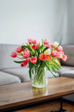 Kahve masasının üzerinde gri kanepe ve grafik yastıklı modern rahat oturma odasının bulanık arka planına sahip taze laleler vazosu. Açık uzay ev iç tasarımı. Uzayı kopyala Dikey kart