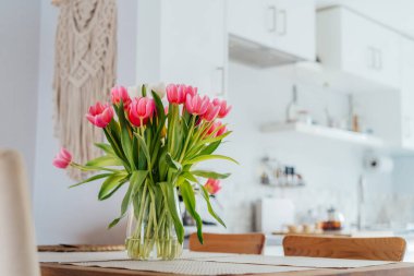 Şık ve modern boho, açık uzay beyaz mutfağının iç mekanı ahşap mutfak masasındaki vazoda pembe lale çiçekleri, duvarda makrome. Samimi Tasarım ev dekorasyonu. Seçici odak.