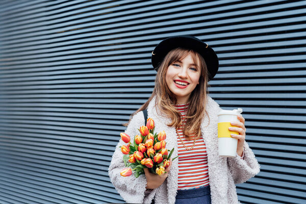 Portrait of happy, smiling young fashion woman in hat holding reusable coffee cup and bouquet of tulip flowers on the gray striped wall background. Eco friendly urban city street fashion. Spring mood