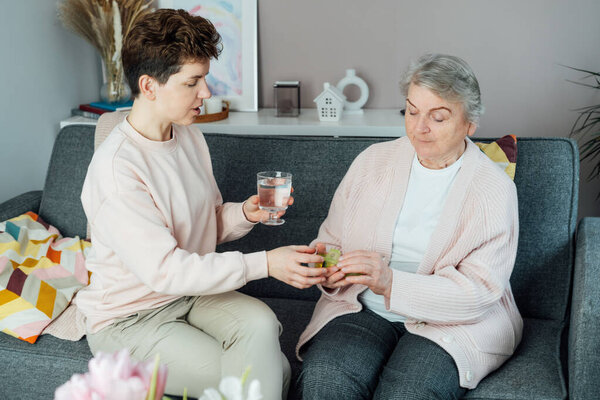 Middle aged neutral gender daughter taking care of old mother, giving pills or vitamin supplements and glass of water helping her mom taking medicine on time. Elderly healthcare and support