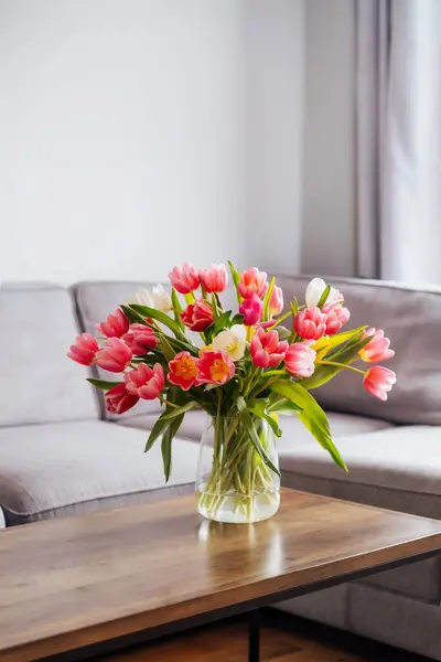 Vase of fresh tulips on the coffee table with blurred background of modern cozy light living room with gray couch sofa and graphic cushions. Open space home interior design. Copy space. Vertical card