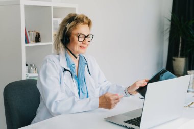 Kulaklıklı kadın doktor bilgisayarında online video görüşmesi yapıyor. Orta yaşlı kadın terapist ev içi sağlık tedavisi için video konferansı yapıyor. Teletıp. Uzaktan kumandalı tıbbi randevu