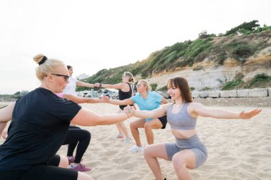 Çeşitli yaşlardaki kadınlar plajda koçla antrenman yapıyor. Hanımlar eşleşen çömelme egzersizleri yapıyor ve birbirlerine sarılıyorlar. Sağlık ve refah için spor. Etkin yaşam biçimi