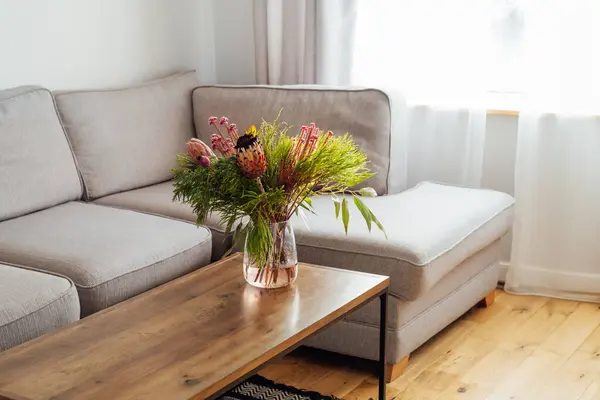エキゾチックなタンパク質の花がコーヒーテーブルに花束を咲かせ モダンな居心地の良いライトリビングルームとグレーのソファーがあります オープンスペースのホームインテリアデザイン エレガントな家の装飾 コピースペース ストックフォト