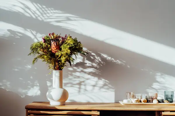 斯堪的纳维亚家庭内部 装饰着异国情调的花束 插在陶瓷花瓶里 站在阳光下的木制橱柜上 阴影笼罩在白色的灰墙上 家居装饰的最低限度设计 免版税图库图片