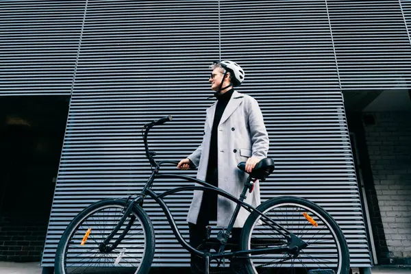 身穿外套 戴着太阳镜 头戴防护头盔 头戴复古自行车的时尚男性站在灰色城墙附近 中性碳足迹运输 绿色生态友好型可持续交通 免版税图库照片