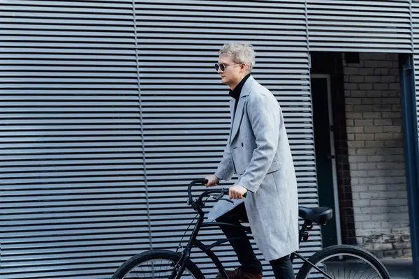 チェックされたコートとサングラスのスタイリッシュな男性は 晴れた日の灰色の都市壁の近くのレトロな自転車で立っています 中立カーボンフットプリント輸送 グリーンエコフレンドリーなモビリティの持続可能な輸送 ストック画像