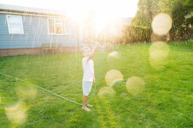 Gülümseyen neşeli çocuk su püskürtme hortumuyla oynuyor, çimenleri suluyor. Çocuklar için yaz bahçesi eğlencesi. Güneşli bir günde arka bahçede su sıçrayan bir çocuk. Mutlu aktif çocukluk. Seçici odak