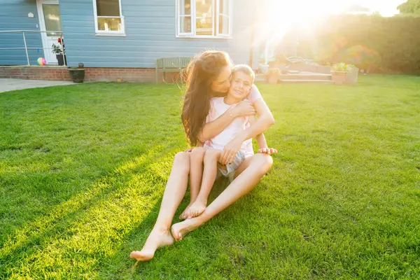 快乐的白人幼儿被快乐的年轻母亲拥抱着 看着相机 面带微笑 夕阳西下 年轻的妈妈和孩子抱着爱 柔情在后院草坪的背景上 图库图片