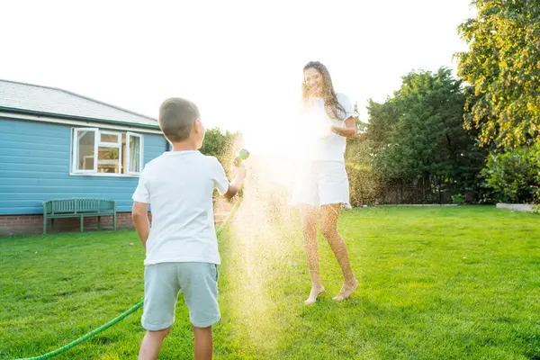 快乐的孩子和妈妈一起玩洒水软管 在日落时一起在花园里玩乐 夏季户外活动 快乐活跃的童年 家庭时光 有选择的重点 图库图片
