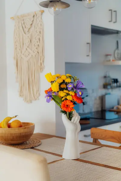 モダンな居心地の良いホワイトキッチンの鮮やかな背景を持つマルチコロールのさまざまな夏の花束と人間の手の形で作られた花瓶に焦点を当てたキッチンカウンターテーブル ホーム インテリアデザインの詳細 ストック写真