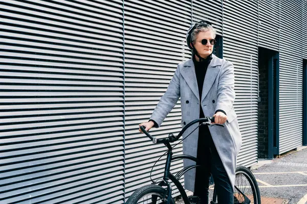 穿着外套 太阳镜和防护头盔的时尚男性骑在灰色城墙附近的复古自行车上 中性碳足迹运输 绿色生态友好型可持续交通 图库照片