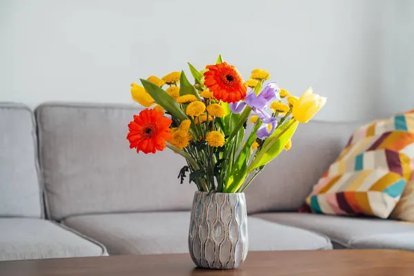 陶瓷花瓶与五彩缤纷的花束在老式风格的木制咖啡桌上 背景模糊的现代舒适轻盈的客厅与灰色沙发沙发 舒适的家居室内设计 图库照片