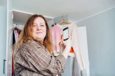 Elbiselerini satmak ya da bağışlamak için akıllı telefonunu kullanan bir kadın kazağın fotoğrafını çekiyor. Temizlik, giysi ayıklama ve gardırop temizleme. Tekrar kullan, ikinci el konsept. Bilinçli tüketici