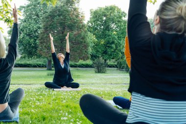 Park 'ta kadın yoga dersi. Bir grup farklı kadın esneme pozu veriyor yeşil çimlerin üzerinde eğitmenle birlikte egzersiz yapıyorlar. Sağlık, refah ve akıl sağlığı için spor faaliyetleri. Seçici odak.