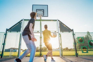 Gün batımında parkın bahçesinde basketbol oynayan iki genç beyaz erkek. Gençlerin hobisi, çevrimdışı aktiviteler. Spor anlayışı, hobiler ve sağlıklı yaşam tarzı. Seçici odak