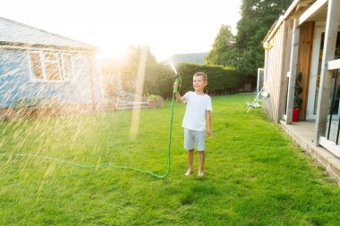 Gülümseyen neşeli çocuk su püskürtme hortumuyla oynuyor, çimenleri suluyor. Çocuklar için yaz bahçesi eğlencesi. Güneşli bir günde arka bahçede su sıçrayan bir çocuk. Mutlu aktif çocukluk. Seçici odak.