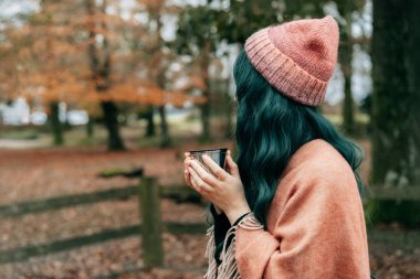 Şallı pançoya sarılı şapkalı kadın sonbahar yürüyüşü sırasında ormanda yapraklarla termos bardağından sıcak içecek içiyor. Uyum, rahatlama, kişisel tatmin. Yerel seyahat yaşam tarzı