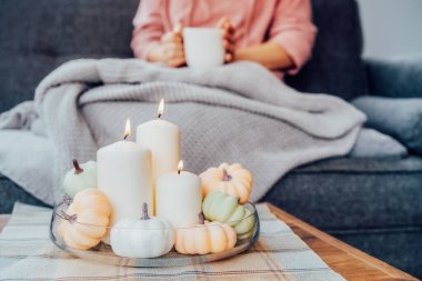 Autumn 'a odaklan, kanepede dinlenen, elinde ekose bir bardak sıcak içecek olan bir kadınla Hygge için rahat bir ortam kompozisyonu oluştur. Dekoratif balkabakları ve sehpada yanan mumlar.