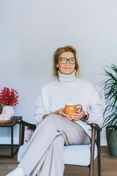 stock image Middle aged woman relaxing with pumpkin shaped cup of hot drink in scandy style hygge interior home with fall mood decor. Lady dreaming, enjoy calm mood without stress, well being alone. Cozy autumn.