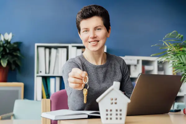 stock image Smiling neutral gender businesswoman real estate agent broker holding house keys with house model miniature, laptop with ready contract for deal with client on table. mortgage or rent concept.