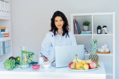 Pozitif Hintli kadın beslenme uzmanı ofis masasında taze sebzelerle müşteriyle görüşüyor, besin takviyeleri ve vitaminler yazıyor, dengeli bir diyet planı yapıyor.