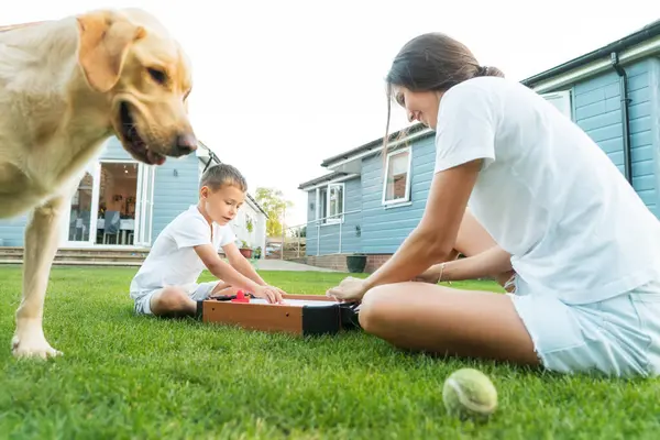 stock image Joyful little boy with mother are playing portable air hockey in garden while dog pet waiting for throwing ball. Fun Playing Games in Backyard Lawn on Summer Day. Happy family time with pet together