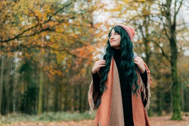 Sıcak şalla kaplanmış şapkalı bir kadın ormanda sonbahar yürüyüşünün tadını çıkarıyor. Seyahat yaşam tarzı. Ahengi hissetmek, doğayla bir araya gelmek. Rahatlatıcı, kişisel tatmin. Yerel seyahat.