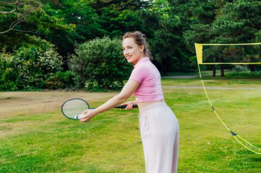 Şehir parkında raketle badminton oynayan gülümseyen genç bir kadın. Arkadaşlar dışarıda eğleniyor. Oyunun ve doğal çevrenin tadını çıkarıyorum. Yetişkin açık hava aktivitesi. Sağlıklı yaşam tarzı