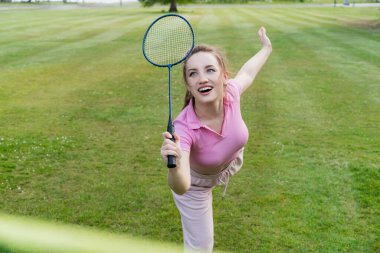 Şehir parkında raketle badminton oynayan gülümseyen genç bir kadın. Arkadaşlar dışarıda eğleniyor. Oyunun ve doğal çevrenin tadını çıkarıyorum. Yetişkin açık hava aktivitesi. Sağlıklı yaşam tarzı