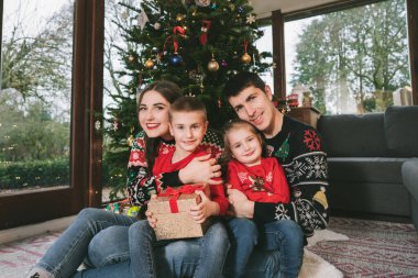 Noel 'de Noel ağacının altında hediyelerle oturmuş neşeli süveterlerle gülümseyen aile fotoğrafı. Mutlu ebeveynler ve çocuklar sarılıyor, heyecanlı hissediyorlar. Ailecek vakit geçireceğiz. Mutlu çocukluk