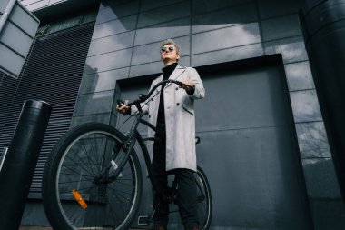 Güneşli bir günde gri kentsel duvarın yanında eski bir bisikletle duran, ceketli ve güneş gözlüklü şık bir erkek. Tarafsız karbon ayak izi taşımacılığı. Yeşil çevre dostu hareketlilik sürdürülebilir ulaşım