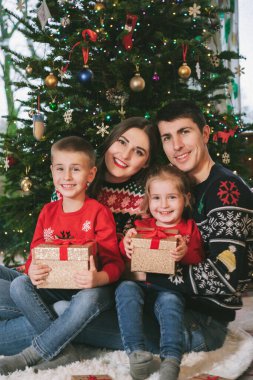 Noel 'de Noel ağacının altında hediyelerle oturmuş neşeli süveterlerle gülümseyen aile fotoğrafı. Mutlu ebeveynler ve çocuklar sarılıyor, heyecanlı hissediyorlar. Ailecek vakit geçireceğiz. Mutlu çocukluk