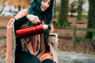 Şallı pançoya sarılı bir kadın sonbahar yürüyüşü sırasında piknik masasına oturup termostan sıcak su döker. Uyum, rahatlama, kişisel tatmin. Yerel seyahat yaşam tarzı