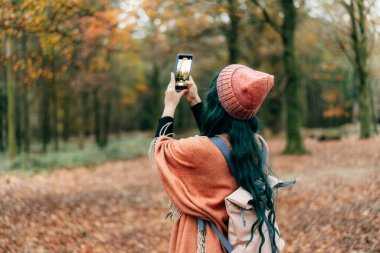 Sırt çantasıyla şallı bir kadın ormanda sonbahar yürüyüşünün tadını çıkarırken telefonla fotoğraf çekiyor. Anı yakala. Uyum hissediyorum. Rahatlatıcı, kişisel tatmin. Yerel seyahat yaşam tarzı.