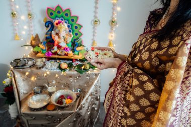 Indian woman dressed in traditional wear engages in worship at home altar, adorned with decors and offerings for god Ganesha during festive celebration. Indian culture, hindu ritual and customs clipart