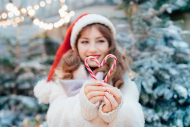 Geleneksel Noel pazarında Noel Baba şapkalı genç bir kadının baston şekerlerinden yapılmış kalp şekline odaklan. Mutlu yıllar Noel tatilleri. Aşkı paylaşın, hayırseverlik konsepti.