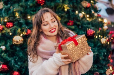 Gülümseyen kadın, geleneksel şehir caddesi Xmas pazarında süslenmiş Noel ağacının arka planında hediye kutusu tutuyor. Hediye değiş tokuşu. Şenlik havası. Festival alışverişi, satış, indirim..