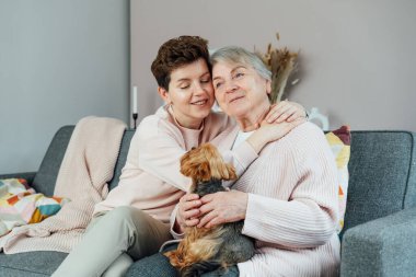 70 'li yaşlarda kızıyla gülümseyen, Yorkshire teriyerinin köpeği koltukta sarılıyor. Aile hafta sonu birlikte. Ebeveyn sevgisi, bakım, nesiller boyu birlik kavramı. Emeklilik evinde kaliteli zaman geçirmek