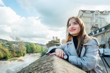 Portrait of Young Woman enjoying sunny day and view of bay with river in picturesque location of Bath city, England. Holiday, travel walk in old European city. Local travel, Sightseeing trip concept clipart