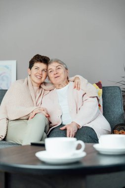 Tarafsız bir cinsiyet kadını, kıdemli annesine ya da büyükannesine sarılıyor ve televizyon seyrediyor, birlikte hassas anlarını paylaşıyor. Ebeveyn sevgisi, bakım ve nesiller boyu birlik kavramı. Anneler Günü. Mutlu aile hafta sonu eğleniyor