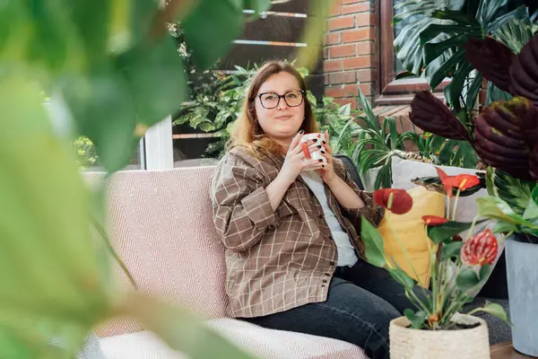 Bir bardak içkiyle gülümseyen kadın ev işleri yaptıktan sonra bahçe konservatuarındaki kanepede dinleniyor. Evde dinlenen rahat kadın portresi. Bahçıvanlık hobisi. Şehir ormanları. Biyofili yaşam tarzı